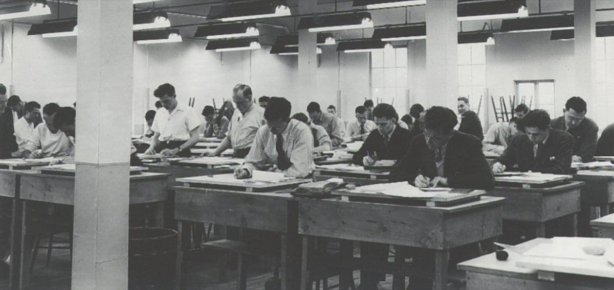 תלמידים יושבים בשורות ליד שולחנות ארוכים בכיתת לימוד בשחור־לבן